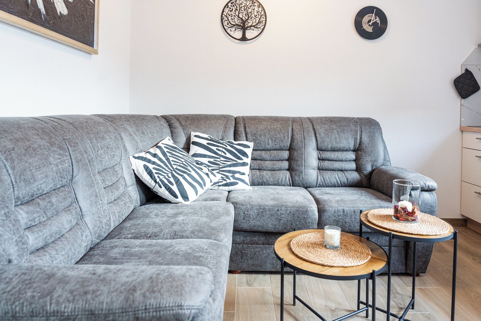 A grey sectional sofa with two round wooden tables, one with a glass and a white object, and a black clock on the wall.
