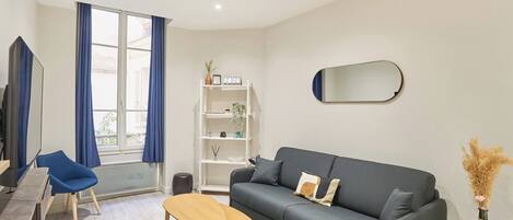 A modern living room with a grey sofa, a wooden coffee table, a blue chair, a white shelving unit, and a large window with blue curtains.