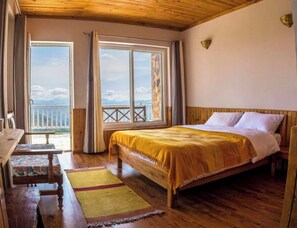 A bedroom with a wooden bed, a bench, and a view of the outdoors through large windows. - Kasar Rainbow Yoga Retreat (Almora)
