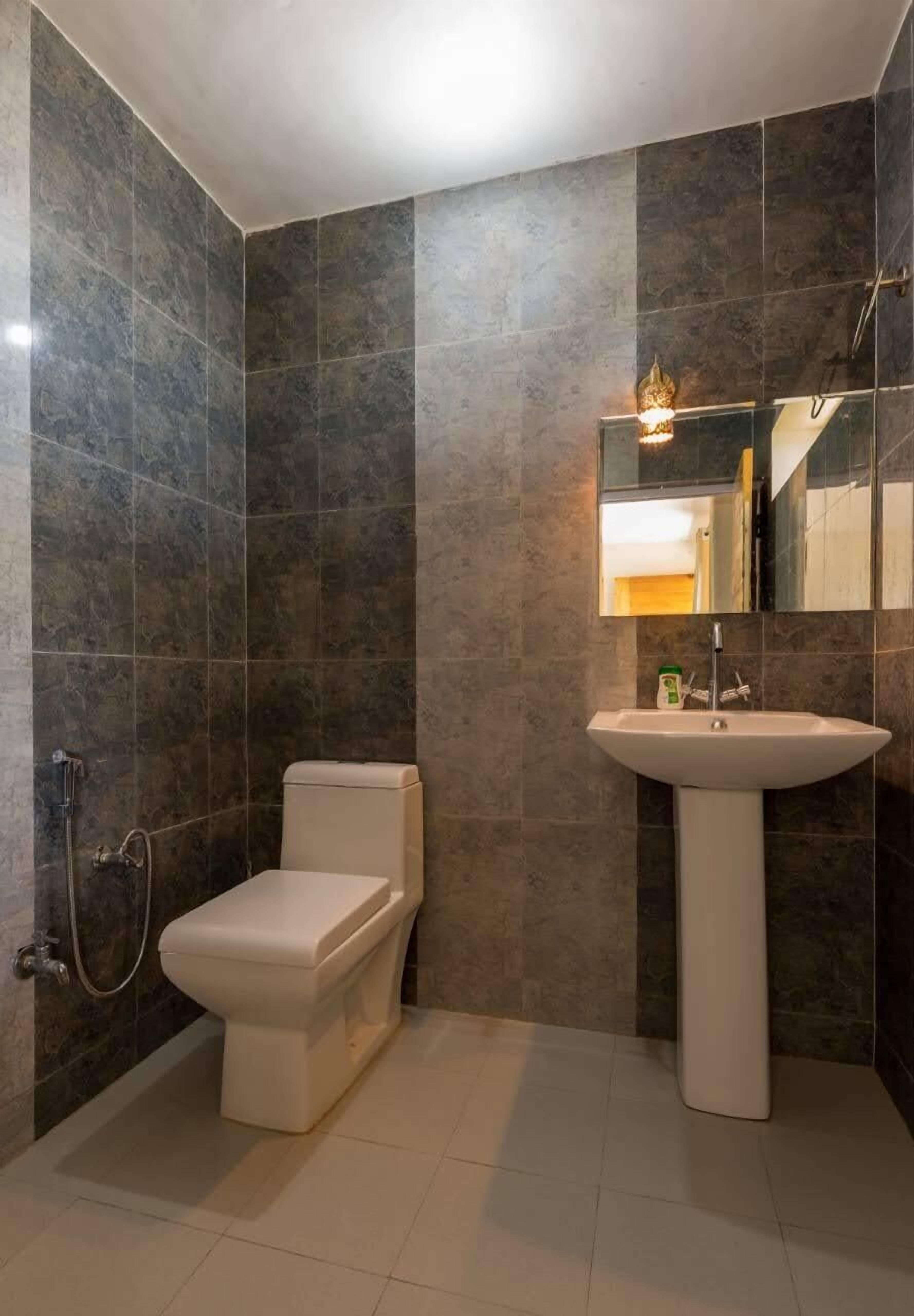 A bathroom with dark tiled walls, a white toilet, a pedestal sink, and a handheld showerhead.