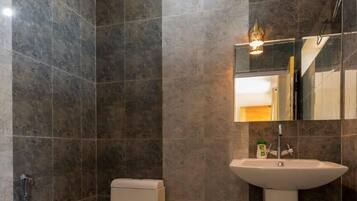 Un baño con paredes de azulejos oscuros, inodoro blanco, lavamanos sobre pedestal y ducha de mano.