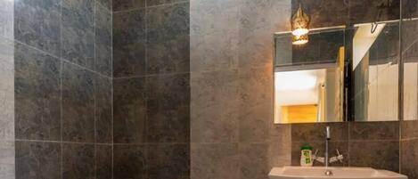 Un baño con paredes de azulejos oscuros, inodoro blanco, lavamanos sobre pedestal y ducha de mano.