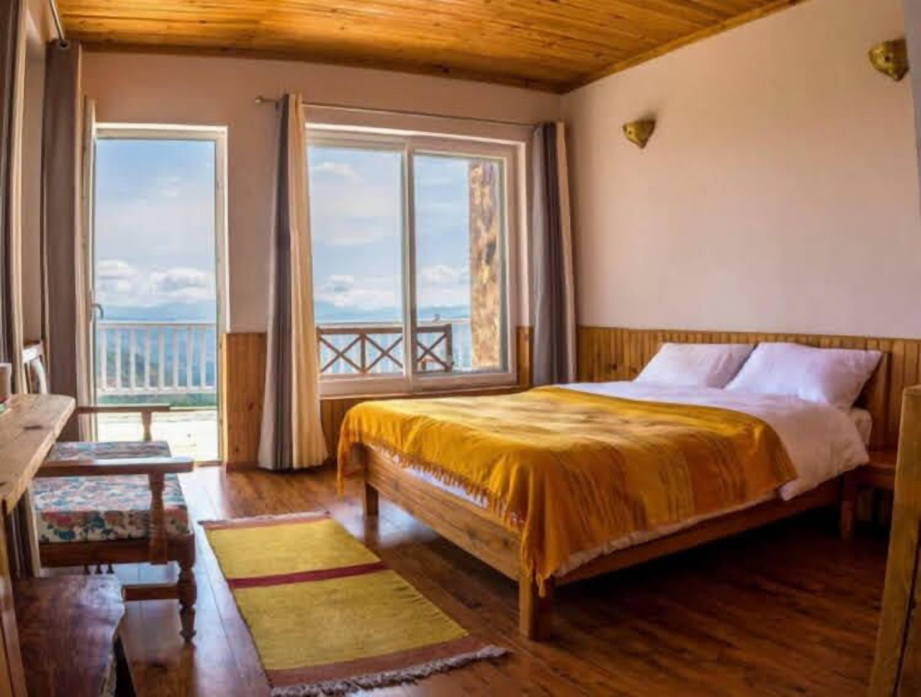 A bedroom with a wooden bed, a desk, and a view of the outdoors through large windows.