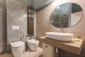 Un baño con pared de mármol, un espejo redondo, un bidé, un inodoro y un lavamanos con encimera color beige.