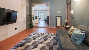 Un salon avec un canapé, un tapis à motifs, un téléviseur fixé au mur et un couloir menant à une autre pièce.