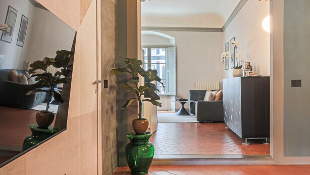 Un couloir orné d’un vase vert, d’une plante en pot et d’un miroir qui reflète une pièce avec un canapé et un radiateur.