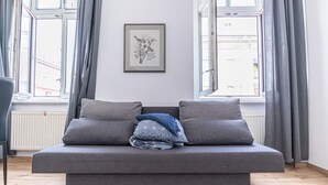 Un saló modern amb un sofà gris, dos coixins, un quadre amb marc a la paret i finestres grans amb cortines.