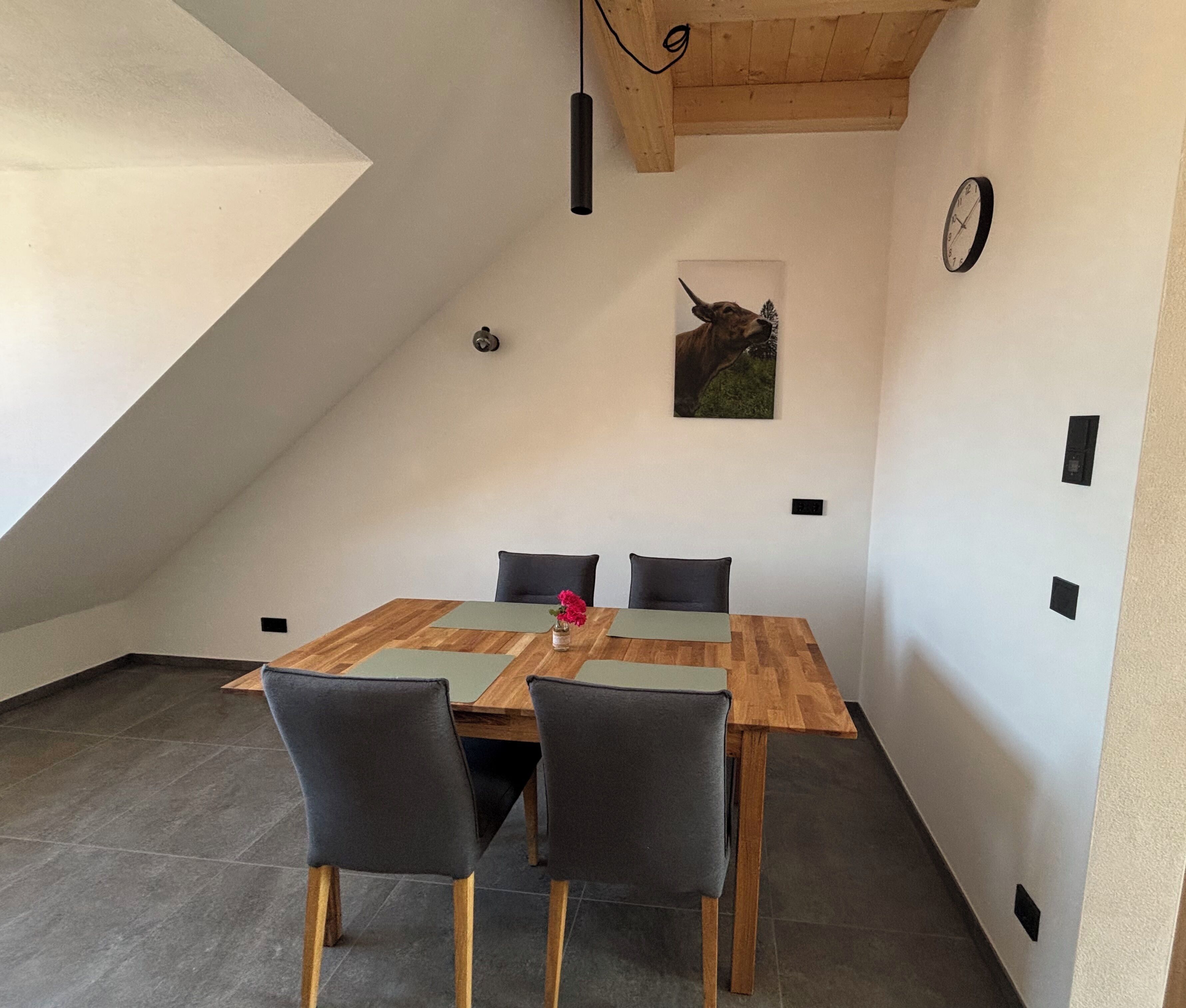 A dining area with a wooden table and four chairs, a clock on the wall, and a ceiling light fixture.