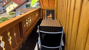A wooden balcony with a table and chairs, a green umbrella, and a view of houses in the background. - NEW: Cozy vacation apartment in Aidling am Riegsee with mountain views (Riegsee)