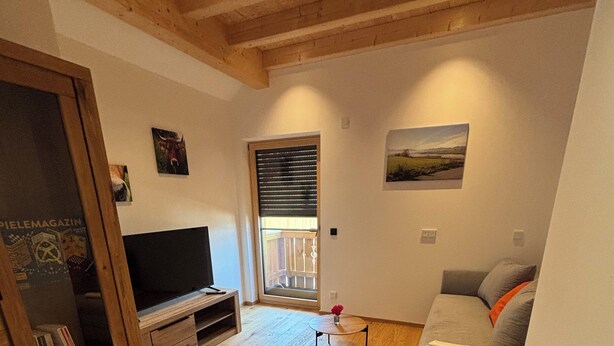 A living room with wooden flooring, a sofa, a TV, a wooden cabinet, and a window with blinds. - NEW: Cozy vacation apartment in Aidling am Riegsee with mountain views (Riegsee)