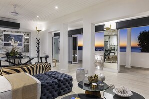 Ein modernes Wohnzimmer mit einem großen Sofa, einem Essbereich und Blick auf den Ozean bei Sonnenuntergang.