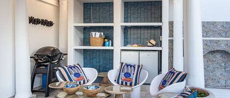 Een moderne buitenruimte met witte stoelen en ronde tafels, een barbecue en een stenen muur.