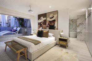 Ein modernes Schlafzimmer mit einem großen Bett, einem Nachttisch, einer Lampe und Blick auf einen Außenbereich mit Pool und Liegestühlen.