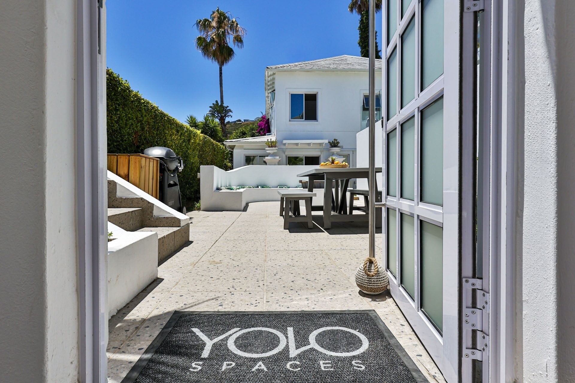 Une terrasse extérieure moderne avec un sol en pierre, un coin salon comprenant une table et des chaises, et un paillasson portant l’inscription « YOLO SPACES ».