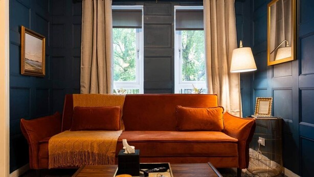 A room with a dark blue paneled wall, a red sofa with a throw blanket, a wooden coffee table with a tray, and a lamp on a side table. - Finnieston 1BR | Walk to Hydro & SEC | Parking (Glasgow)