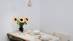 Une table à manger recouverte d’une nappe blanche, deux chaises vertes, un vase avec des tournesols et des lampes suspendues.