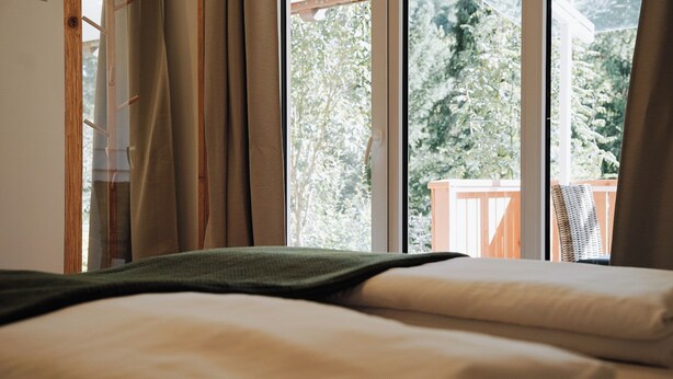 Un lit recouvert d’une couverture verte, une fenêtre avec des rideaux et une vue sur des arbres et une terrasse en bois à l’extérieur.