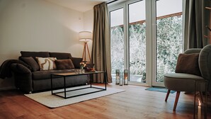 Un salon moderne avec un canapé, une table basse et un fauteuil. Une grande fenêtre, ornée de rideaux, offre une vue sur les arbres à l’extérieur.