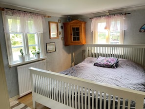 A bedroom with a white crib, a bed with a floral pillow, two windows with sheer curtains, and a radiator. - 5 Person Holiday Home in Immeln (Immeln)