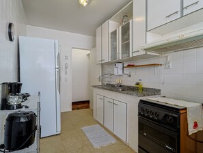 A compact kitchen with white cabinets, a stove, a coffee maker, and a vacuum cleaner. - Apto a 700m da Praia do Leblon com Wi-fi e ar (Rio de Janeiro)