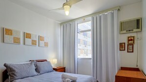 A hotel room with a bed, a desk, a window with curtains, a ceiling fan, and wall art. - Apto a 700m da Praia do Leblon com Wi-fi e ar (Rio de Janeiro)