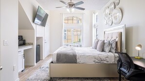 A bedroom with a bed, a desk, a chair, a ceiling fan, and a TV mounted on the wall.