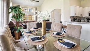 A modern living room with a glass dining table set for four, surrounded by comfortable sofas and a large potted plant.