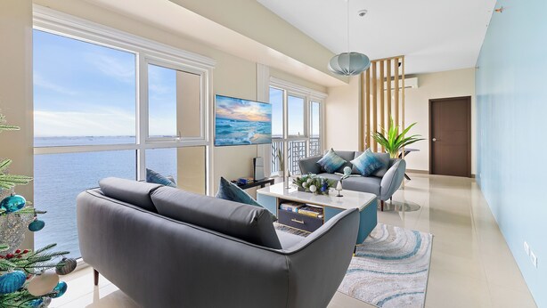 A modern living room with a large window overlooking the sea, a grey sofa, a coffee table, and a potted plant. - All Inclusive VIP Celeb 5 Star Resort Pool Spa Gym (Parañaque)