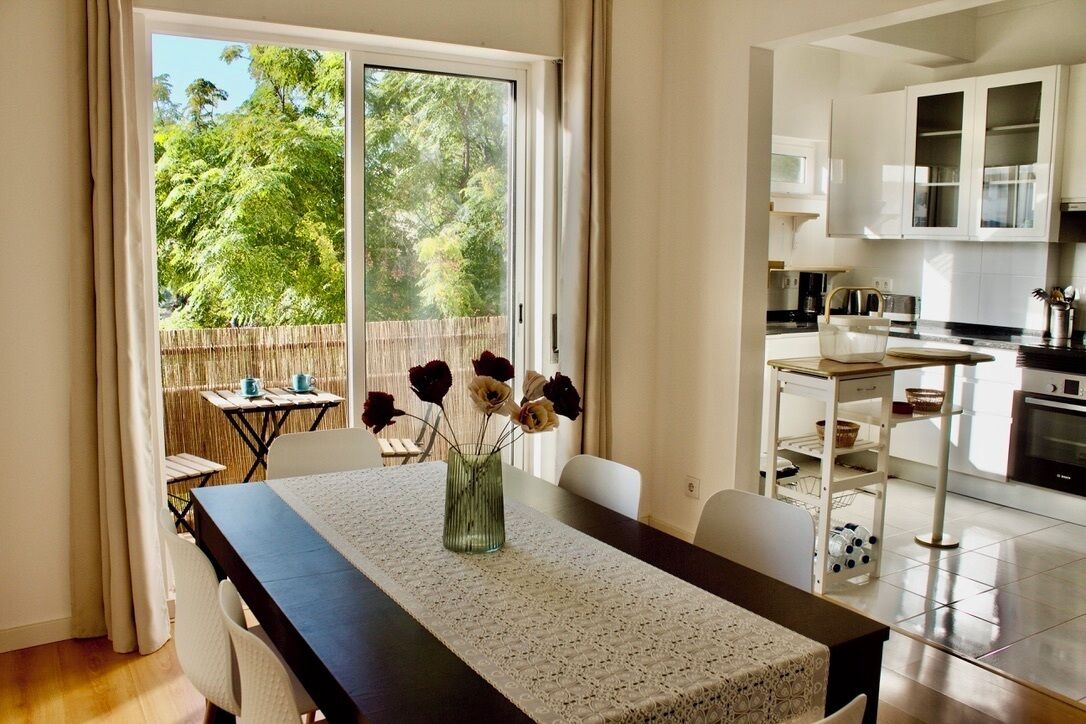 Una cocina moderna con armarios blancos, un comedor con mesa dispuesta para dos personas, y un ventanal con vistas a árboles.