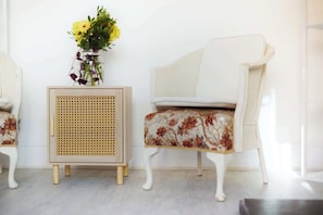 Une chaise blanche recouverte d’un tissu à motifs floraux, une petite table d’appoint en osier avec un vase de fleurs et un mur uni.