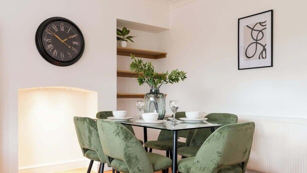 A dining area with a table set for four, green upholstered chairs, a vase with green foliage, a wall clock, and a framed abstract artwork. - Spacious Home | Driveway Parking | Minutes from M1 (Nottingham)