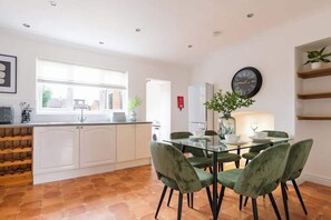 A modern kitchen with white cabinets, a wine rack, a dining area with a glass table and green chairs, a clock on the wall, and a window with blinds. - Spacious Home | Driveway Parking | Minutes from M1 (Nottingham)