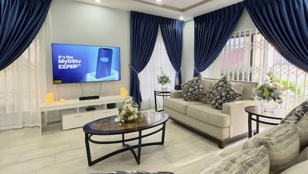A modern living room with a sofa set, a coffee table, and a TV displaying an advertisement. - Spacious 2BR private apartment setting for families near Accra. (Oyibi)