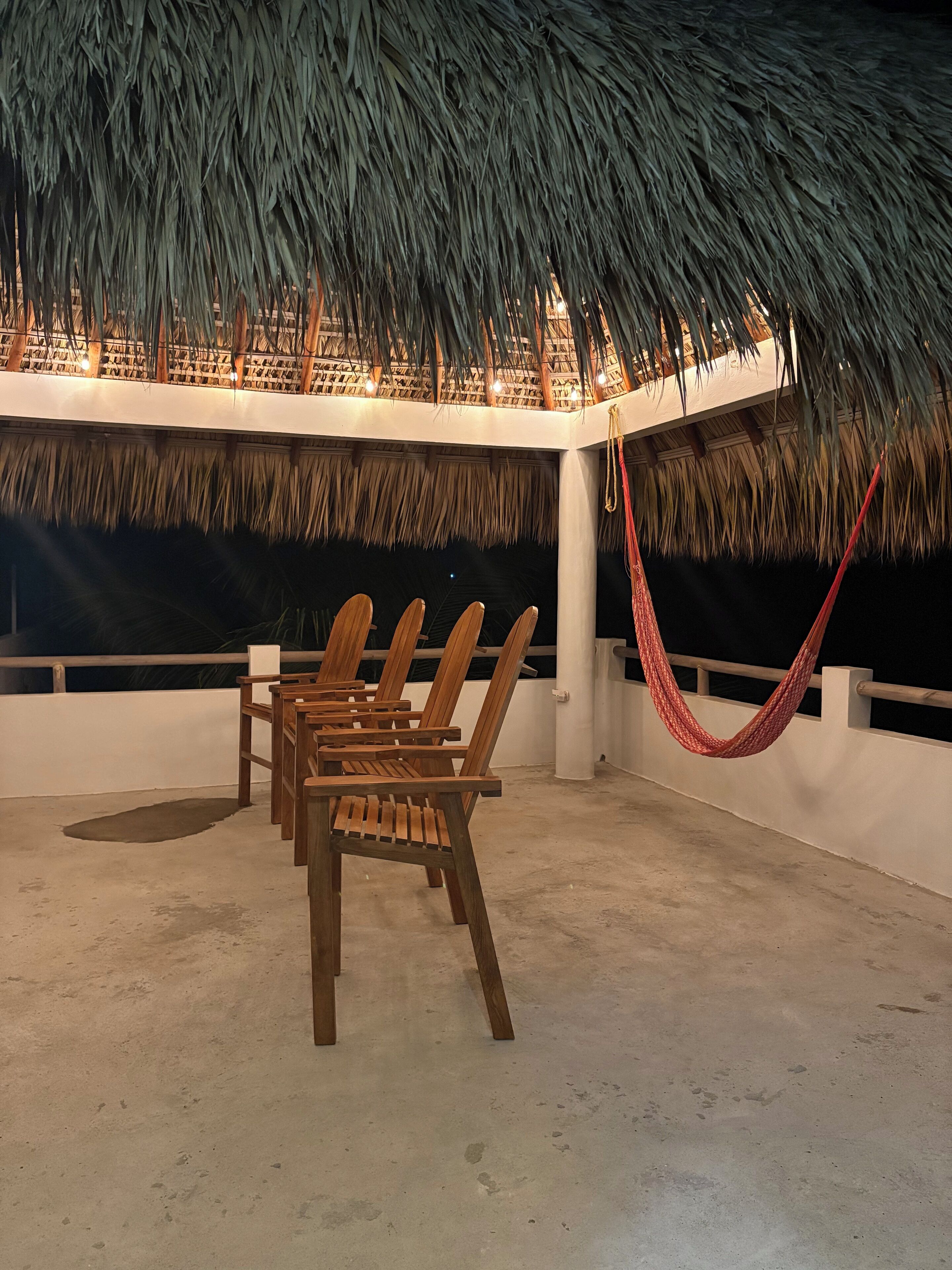 A thatched-roof outdoor seating area with wooden chairs and a red hammock.