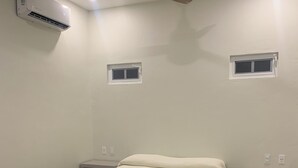 Une chambre à coucher comprenant un lit, deux tables de chevet, un ventilateur de plafond, un appareil de climatisation et deux petites fenêtres.