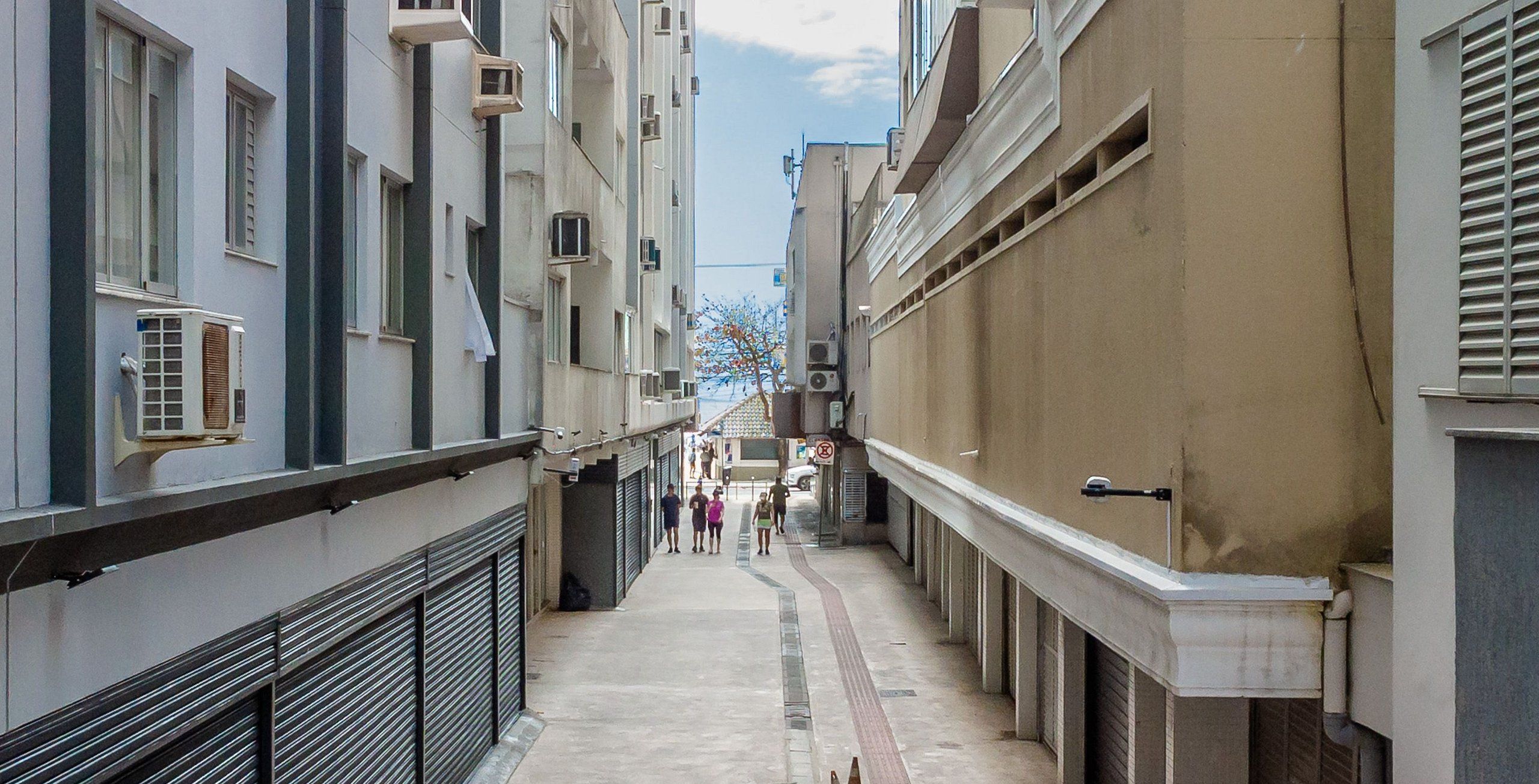 A narrow urban alleyway with buildings on either side, featuring air conditioning units, a visible staircase, and a pedestrian walkway.