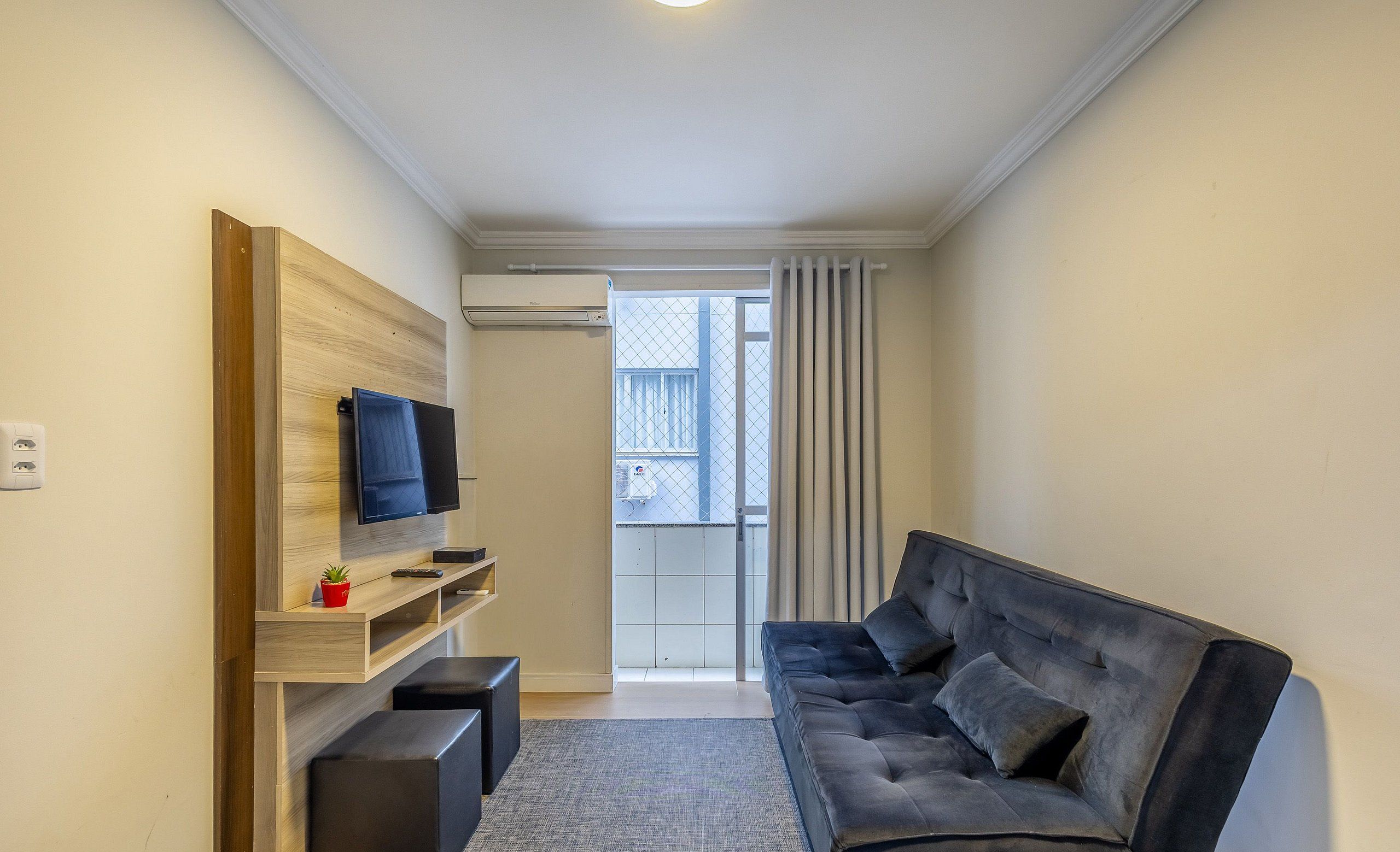 A modern living room with a black sofa, a flat-screen TV mounted on a wooden shelf, and a sliding glass door leading to a balcony.