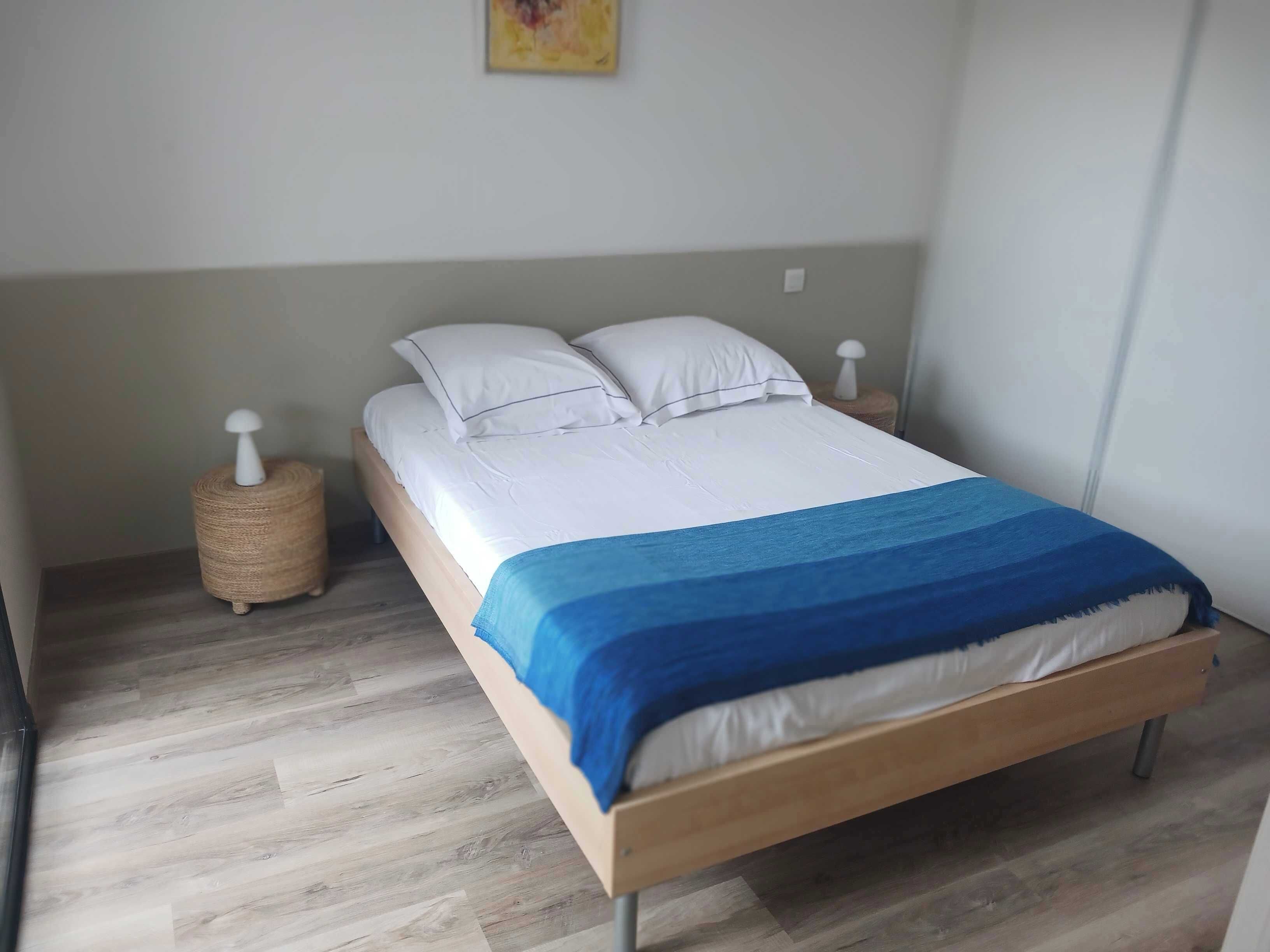 Een net opgemaakt bed met witte lakens en een blauwe deken, een houten hoofdbord, en een klein rond rieten mandje op de vloer naast het bed.
