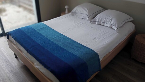 Una cama con colchón blanco y manta azul, cabecera de madera y suelo de madera.