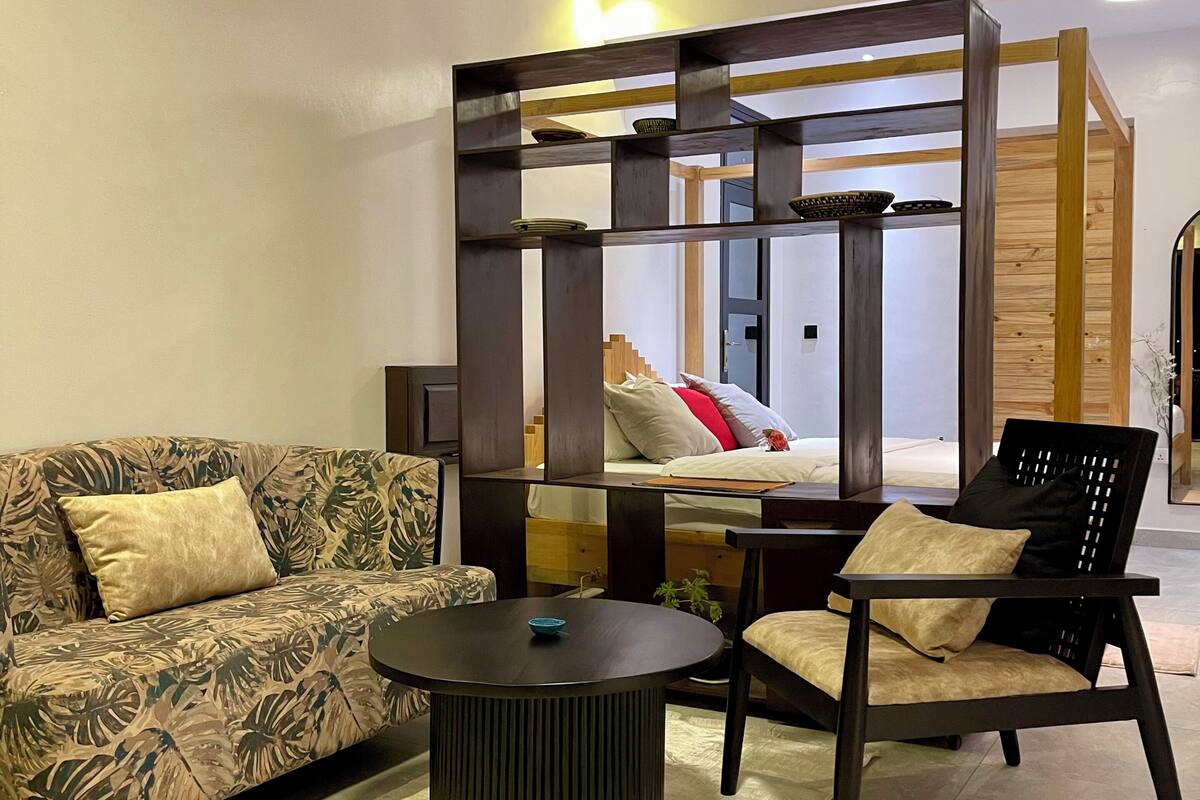 A modern living room with a sofa, a coffee table, and a chair. There is a wooden partition with a mirror and a bed in the background.