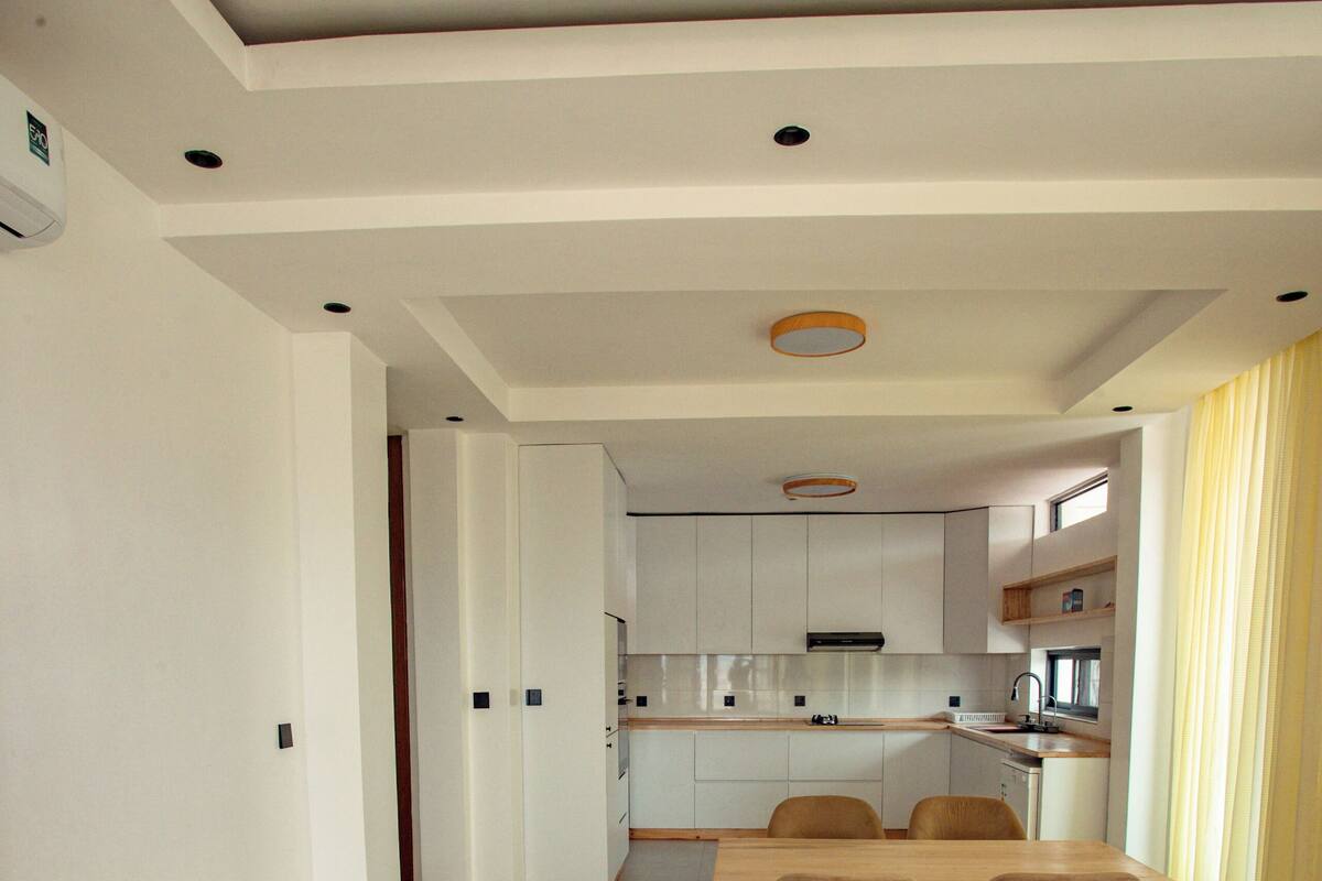 A modern kitchen with white cabinets, a dining area with a wooden table and chairs, and a yellow curtain in the background.