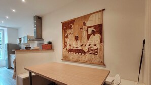 A modern kitchen with a wooden table and chairs, a hanging tapestry depicting traditional figures, and a visible kitchen counter with appliances. - Bright apartment with private beach access (Saint-Lunaire)