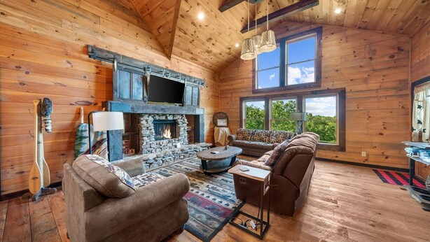 A cozy cabin interior with a stone fireplace, wooden walls, a sofa, and a large window offering a view of the outdoors.