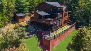 A multi-story log cabin with a balcony, surrounded by a well-maintained lawn and dense forest.