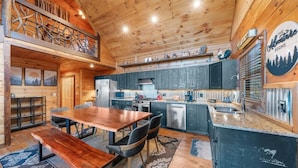 A cozy cabin interior with a wooden dining table, chairs, a kitchen with modern appliances, and a loft area with a view of the mountains.