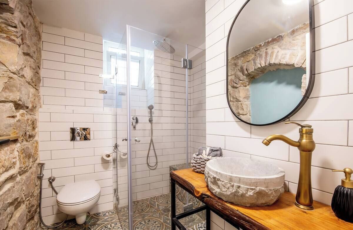 Ein Badezimmer mit weißen Fliesen, einer Akzentwand aus Stein, einem runden Spiegel, einer Holzwaschbeckenbank, einem Steingefäß, einem Messingarmaturen und einer Toilette.