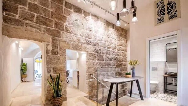 Un interior moderno con una pared de piedra, una zona de comedor con mesa y sillas, y una cocina visible a través de una puerta abierta.