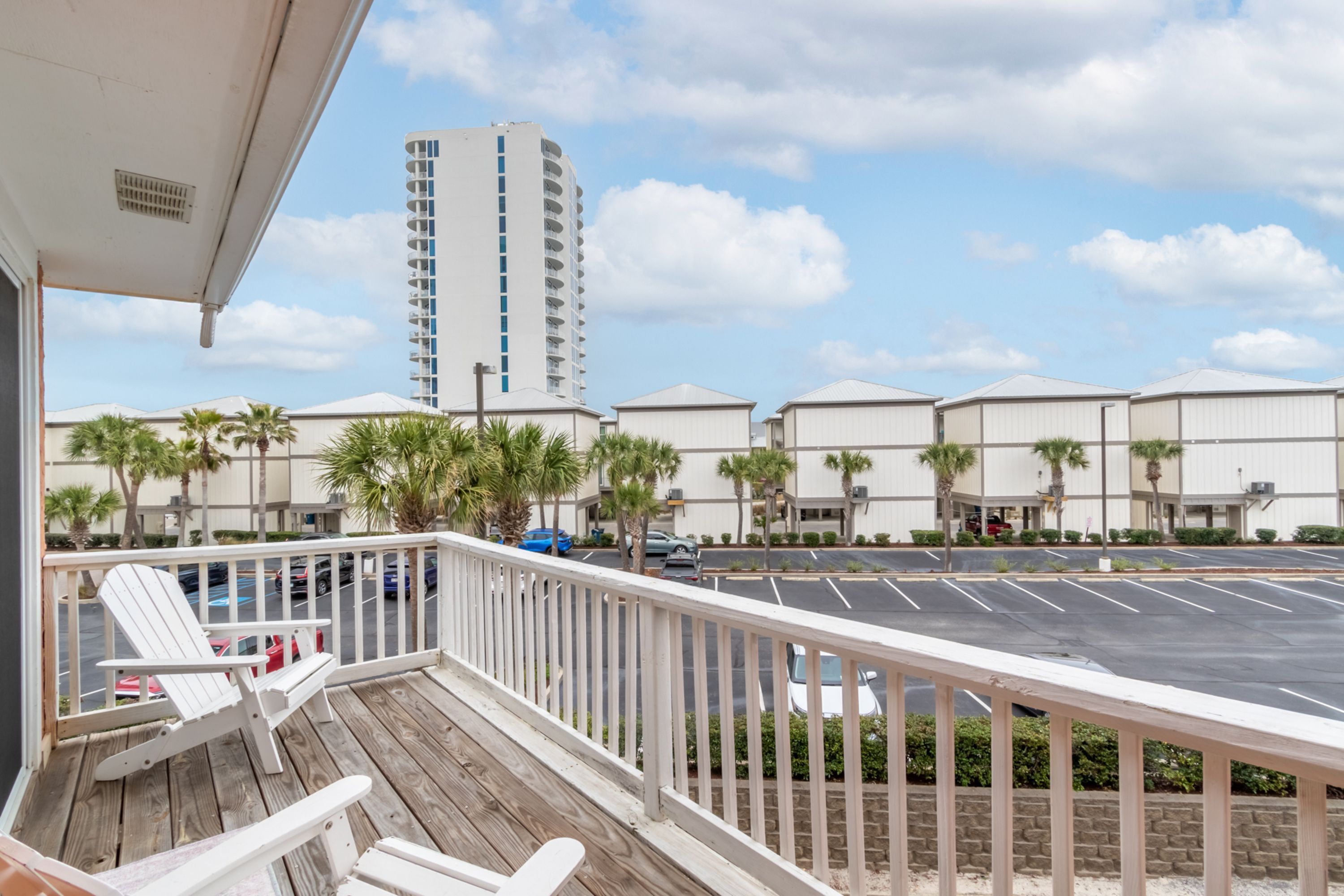 A wooden deck with two Adirondack chairs, a view of a parking lot, and a tall building in the background.