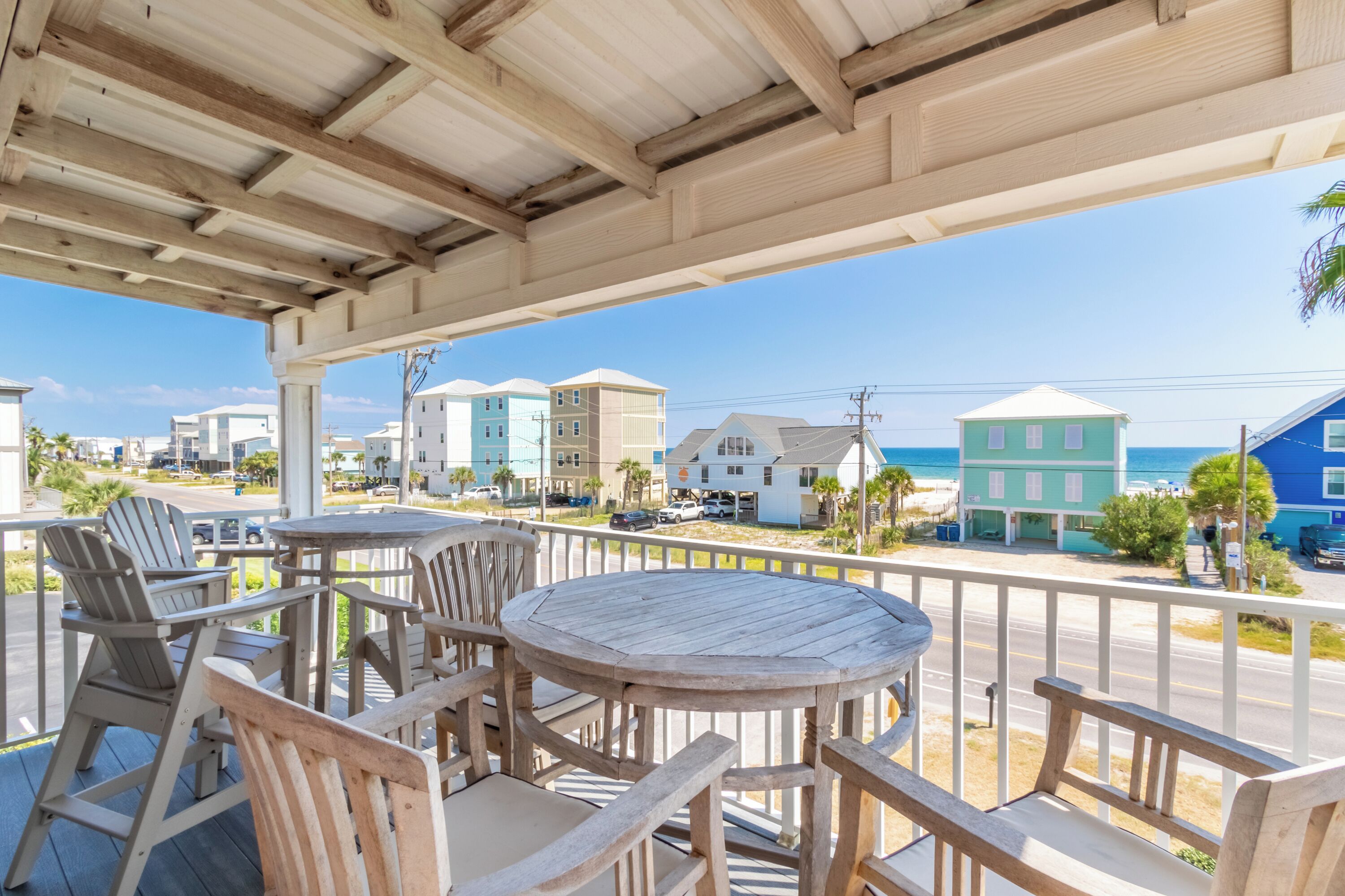 Une terrasse en bois avec des sièges extérieurs, offrant une vue sur une plage bordée de bâtiments aux couleurs vives et un ciel dégagé.
