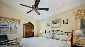 Un dormitorio con cama, ventilador de techo, armario de madera, escritorio y ventana con cortinas.